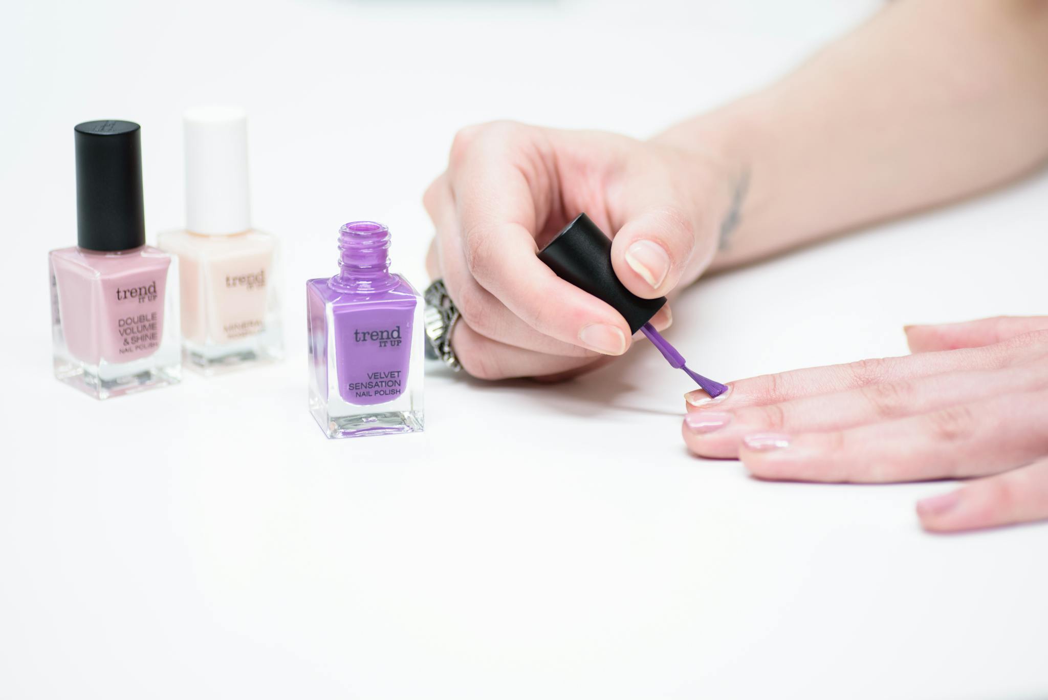 A woman applying purple nail polish with bottles of various colors in the background.