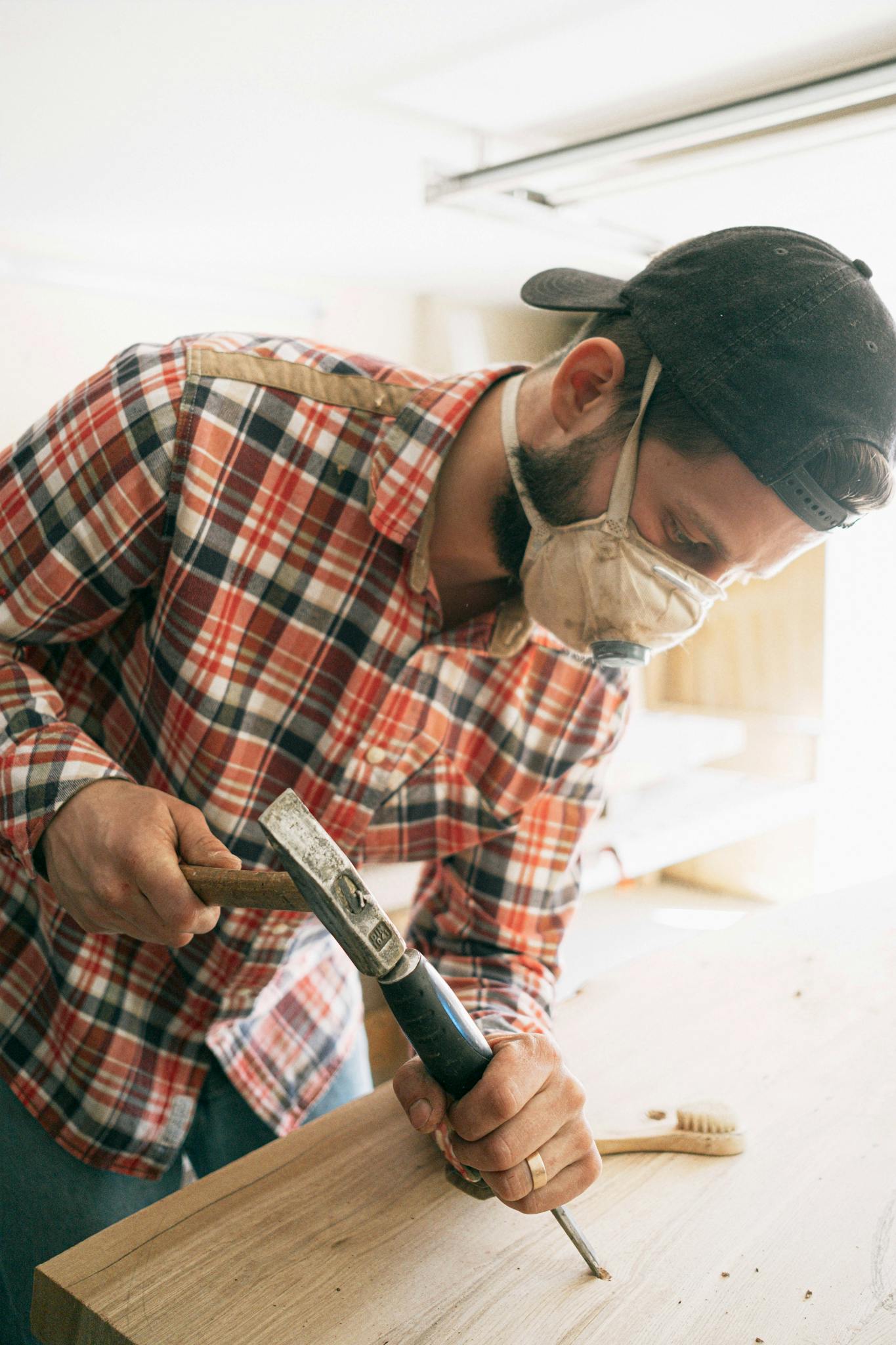 Focused carpenter working with tools in a sunlit workshop, wearing a face mask for safety.